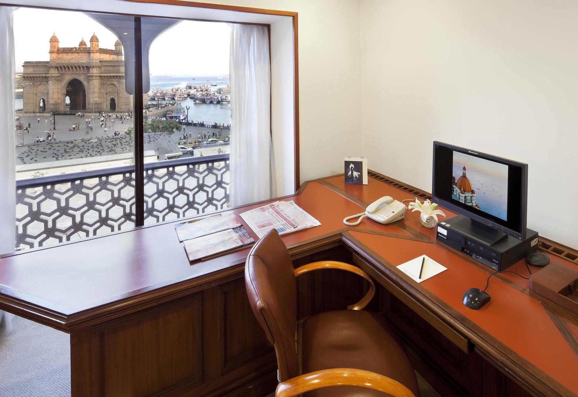 فندق ذا تاج محل تاور مومباي المظهر الخارجي الصورة The photo shows a workspace with a wooden desk and an ergonomic chair. On the desk, there is a computer monitor, a telephone, and some stationery items. A window provides a view of an outdoor scene, possibly featuring water and structures or building